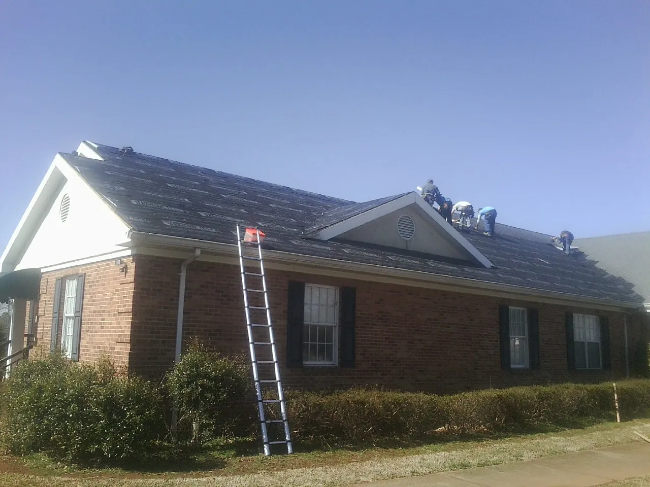 Crew tearing shingles off a brick ranch roof with a ladder set — Emergency Roof Repair work by our crew in Gallitzin
