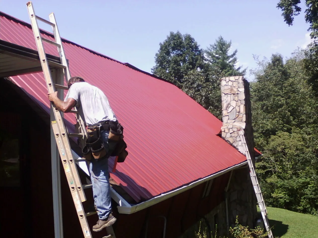 Roofer on a ladder beside a red metal roof and stone chimney — Soffit & Fascia Repair by Roofing Brothers in Gallitzin